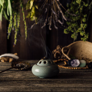 Brûle-parfum en céramique fumant sur une table en bois, bouquets d’herbes séchées derrière